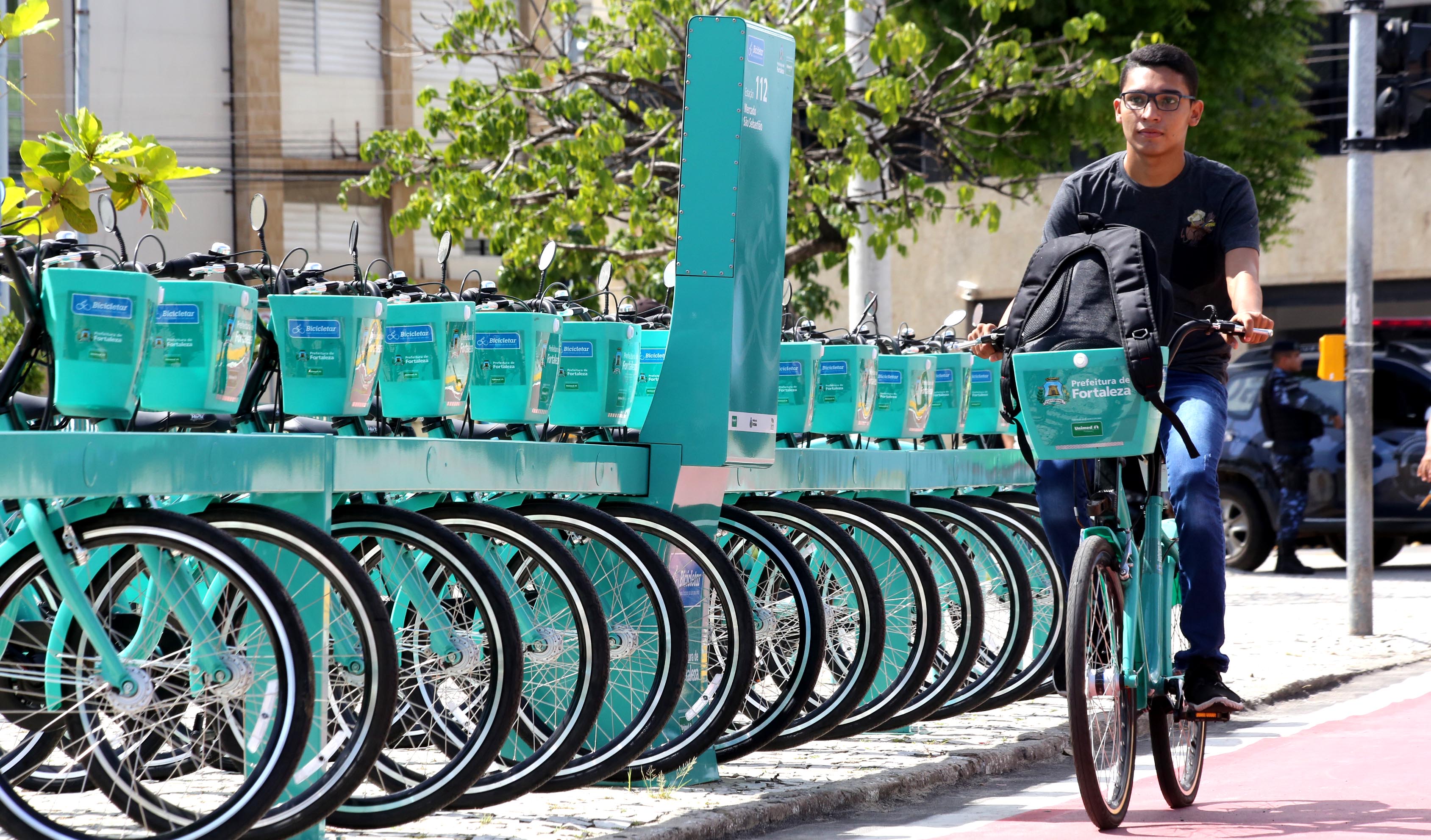 jovem andando de bibicleta em ciclofaixa ao lado de estação com várias bicicletas estacionadas